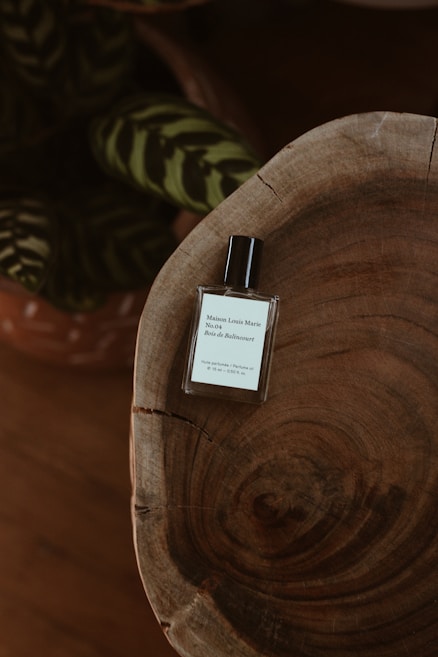A minimalist and elegant perfume bottle sits atop a rustic wooden surface with visible grain patterns. In the background, there are dark green leaves with lighter stripes, adding a touch of nature to the composition.