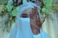 A person wearing a white veil and a gem-studded tiara, with one hand adorned with intricate henna designs raised to cover part of their face. The background features soft-focus flowers in light green and white hues.