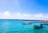 Several small boats are floating on a bright turquoise sea under a clear blue sky with scattered clouds. A distant coastline with buildings and a small pier are visible in the background.