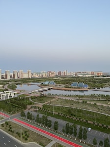 A serene urban landscape featuring high-rise buildings in the background, surrounded by lush greenery and calm water bodies. Solar panels are visible on the banks, indicating a focus on eco-friendly solutions. The area includes paved walkways and well-maintained roads, enhancing accessibility amidst a blend of natural and urban elements.