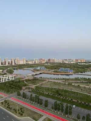 A serene urban landscape featuring high-rise buildings in the background, surrounded by lush greenery and calm water bodies. Solar panels are visible on the banks, indicating a focus on eco-friendly solutions. The area includes paved walkways and well-maintained roads, enhancing accessibility amidst a blend of natural and urban elements.