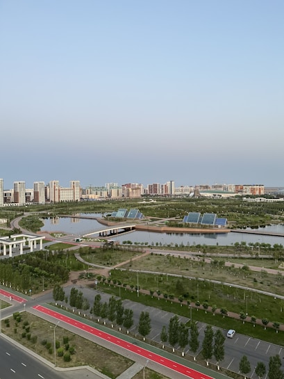 A serene urban landscape featuring high-rise buildings in the background, surrounded by lush greenery and calm water bodies. Solar panels are visible on the banks, indicating a focus on eco-friendly solutions. The area includes paved walkways and well-maintained roads, enhancing accessibility amidst a blend of natural and urban elements.