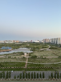 A sprawling urban landscape with a mix of natural and constructed elements. The foreground features a parking lot and neatly arranged trees lining the area. In the midground, there are extensive green spaces, including manicured lawns, small ponds, and pathways. The background is dominated by clusters of high-rise buildings and a large structure resembling a stadium. The sky above is clear with a gradient from blue to light at the horizon.