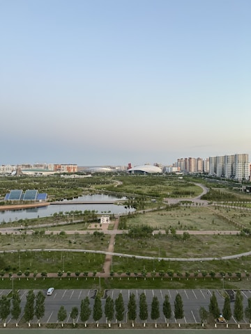 A sprawling urban landscape with a mix of natural and constructed elements. The foreground features a parking lot and neatly arranged trees lining the area. In the midground, there are extensive green spaces, including manicured lawns, small ponds, and pathways. The background is dominated by clusters of high-rise buildings and a large structure resembling a stadium. The sky above is clear with a gradient from blue to light at the horizon.