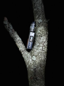 A vape device is positioned in the fork of a tree branch against a dark background. The device has a digital display and buttons on the front.