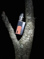 A vape device rests between the branches of a tree, surrounded by bark and lichen in a dark setting.