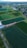 Aerial view of an expansive agricultural area divided into large rectangular plots of farmland. These plots are various shades of green, indicating different types of crops or stages of growth. Adjacent to the farmland is a small residential area with houses and trees, and there are some buildings and structures near the fields. In the background, a small body of water is visible as well as a distant mountain range. The layout of the farmland is organized and there are several dirt roads and paths intersecting the fields.