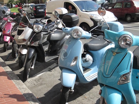 A row of parked scooters in various colors, including pink, black, silver, and light blue, lined up on a street with several cars in the background. The scooters are tightly packed together and situated near a curb paved with red bricks, indicating an urban environment.