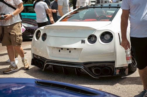A white sports car is parked with people standing around it, some holding cameras and water bottles. The car features a distinct rear design with four tailpipes and circular tail lights. The scene appears to be outdoors in a sunny environment.