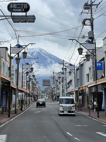 A street in a Japanese town with overhead wires and street signs, featuring a clear view of a snow-capped mountain in the background. The street is lined with traditional and modern buildings, and a few cars are driving along the empty road.
