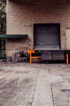 A brick industrial building with a closed metal roll-up door, set on a dirty concrete loading dock area. A small metal awning with a green roof is situated on the left side of the door.