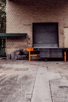 A brick industrial building with a closed metal roll-up door, set on a dirty concrete loading dock area. A small metal awning with a green roof is situated on the left side of the door.
