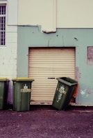 Three green garbage bins with yellow lids are positioned against a wall. Two bins stand upright while the third is slightly tipped over. The wall behind them is painted in contrasting colors with a large closed roller door in the middle. There are some scuff marks and minor graffiti on the wall, including a small heart-like drawing.
