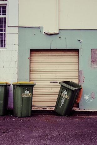 Three green garbage bins with yellow lids are positioned against a wall. Two bins stand upright while the third is slightly tipped over. The wall behind them is painted in contrasting colors with a large closed roller door in the middle. There are some scuff marks and minor graffiti on the wall, including a small heart-like drawing.