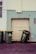 Three green garbage bins with yellow lids are positioned against a wall. Two bins stand upright while the third is slightly tipped over. The wall behind them is painted in contrasting colors with a large closed roller door in the middle. There are some scuff marks and minor graffiti on the wall, including a small heart-like drawing.