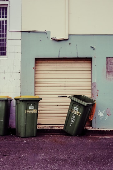 Three green garbage bins with yellow lids are positioned against a wall. Two bins stand upright while the third is slightly tipped over. The wall behind them is painted in contrasting colors with a large closed roller door in the middle. There are some scuff marks and minor graffiti on the wall, including a small heart-like drawing.