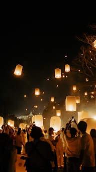 A large group of people gathered under the night sky, releasing floating lanterns that glow warmly as they rise. The lanterns illuminate the scene with a soft, golden light, contrasting against the dark backdrop. Many attendees are taking photos or filming the event, capturing the magical atmosphere. Trees and a distant building can be seen in the background.