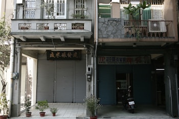 Two adjacent buildings with closed roller shutters. The facade includes some vegetation, potted plants, and signs with Chinese characters. A scooter is parked in front of one building.