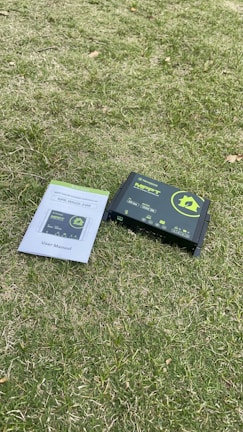 A solar charge controller and its user manual are placed on a grassy surface. The device is primarily black with green and yellow labeling, while the user manual is white with blue and green lettering.