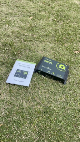 A solar charge controller and its user manual are placed on a grassy surface. The device is primarily black with green and yellow labeling, while the user manual is white with blue and green lettering.