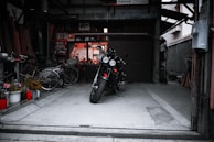 A motorcycle is parked in the center of a dimly lit garage with various items scattered around, including bicycles and potted plants. The space appears cluttered yet organized, with shelves and tools visible in the background.