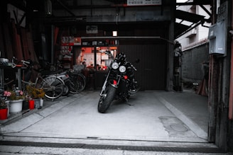 A motorcycle is parked in the center of a dimly lit garage with various items scattered around, including bicycles and potted plants. The space appears cluttered yet organized, with shelves and tools visible in the background.