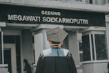 A person wearing a graduation gown and cap is standing in front of a building with the sign 'Gedung Megawati Soekarnoputri'. The image is taken from behind the person, showing the back of their graduation attire. The building appears to be institutional, with grey walls and a somber appearance.