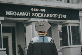 A person wearing a graduation gown and cap is standing in front of a building with the sign 'Gedung Megawati Soekarnoputri'. The image is taken from behind the person, showing the back of their graduation attire. The building appears to be institutional, with grey walls and a somber appearance.