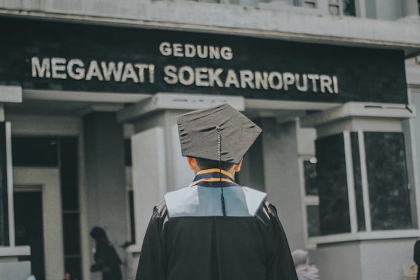 A person wearing a graduation gown and cap is standing in front of a building with the sign 'Gedung Megawati Soekarnoputri'. The image is taken from behind the person, showing the back of their graduation attire. The building appears to be institutional, with grey walls and a somber appearance.