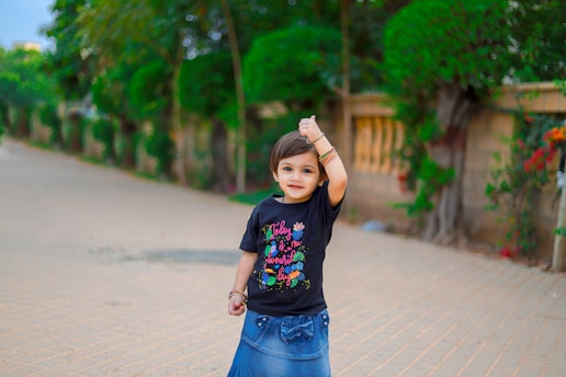 A young child stands on a paved pathway, wearing a black t-shirt with colorful text and graphics. The background features green, manicured trees and a brick wall with flowers.