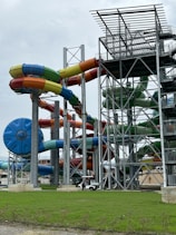 A complex structure of large, colorful water slides intertwined around tall metal supports. The slides are predominantly blue, green, yellow, and red, and are part of a water park station. The ground is covered with lush green grass, and there is an overcast sky in the background.