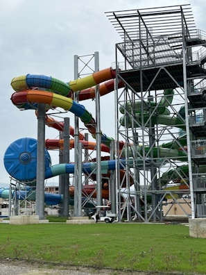 A complex structure of large, colorful water slides intertwined around tall metal supports. The slides are predominantly blue, green, yellow, and red, and are part of a water park station. The ground is covered with lush green grass, and there is an overcast sky in the background.