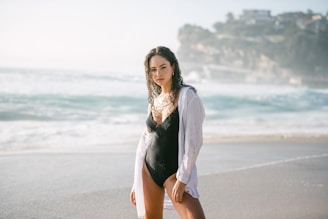 A woman stands on a sandy beach with ocean waves in the background. She is wearing a black swimsuit and a white shirt open over it. The setting appears to be bright and sunny, with cliffs or buildings visible in the distance.