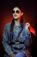 A person wearing round sunglasses poses confidently while dressed in a denim jacket with a unique belt featuring a small decorative element. They hold a red handbag over one shoulder. The backdrop is dark, with a warm, red-orange light creating a striking contrast.