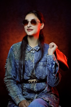 A person wearing round sunglasses poses confidently while dressed in a denim jacket with a unique belt featuring a small decorative element. They hold a red handbag over one shoulder. The backdrop is dark, with a warm, red-orange light creating a striking contrast.