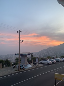 A scenic view captures a city at dusk, with a beautiful gradient sky blending soft pink and orange hues into the blue. The foreground features a road lined with parked vehicles, including cars and motorcycles. A sign for 'Teras Cafe' is visible near a utility pole, and beyond, a sprawling urban landscape extends towards the hills.