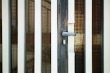 An iron gate with vertical bars and a metal mesh screen is prominently featured. The gate includes a handle and a keyhole at the center, suggesting it is a door or entrance. The metal bars are painted white and have a slightly worn appearance, suggesting some age or weathering.