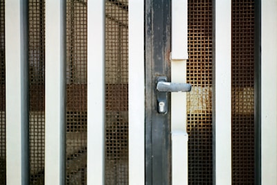 An iron gate with vertical bars and a metal mesh screen is prominently featured. The gate includes a handle and a keyhole at the center, suggesting it is a door or entrance. The metal bars are painted white and have a slightly worn appearance, suggesting some age or weathering.