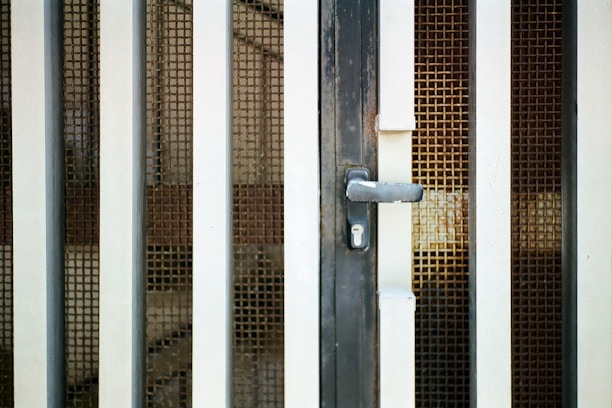 An iron gate with vertical bars and a metal mesh screen is prominently featured. The gate includes a handle and a keyhole at the center, suggesting it is a door or entrance. The metal bars are painted white and have a slightly worn appearance, suggesting some age or weathering.