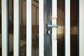 An iron gate with vertical bars and a metal mesh screen is prominently featured. The gate includes a handle and a keyhole at the center, suggesting it is a door or entrance. The metal bars are painted white and have a slightly worn appearance, suggesting some age or weathering.