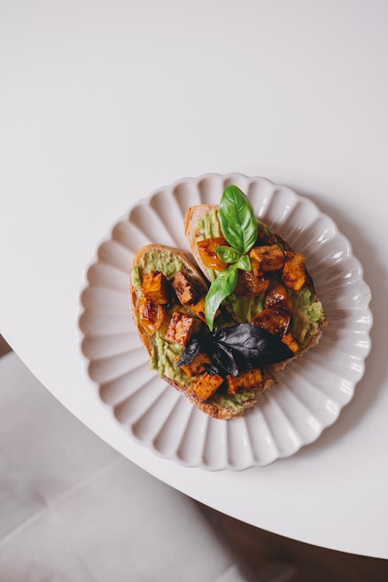 A white ceramic plate with scalloped edges holds a slice of bread topped with mashed avocado, roasted sweet potato cubes, and garnished with fresh basil leaves. The plate is placed on a white table, highlighting the colorful and appetizing presentation of the food.