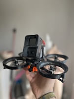 A hand is holding a small drone equipped with a camera at the front. The drone has four propellers, and the camera is enclosed within a black casing. The background is softly blurred, emphasizing the focus on the drone.