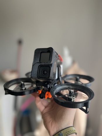 A hand is holding a small drone equipped with a camera at the front. The drone has four propellers, and the camera is enclosed within a black casing. The background is softly blurred, emphasizing the focus on the drone.