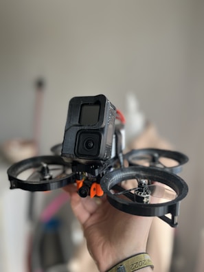 A hand is holding a small drone equipped with a camera at the front. The drone has four propellers, and the camera is enclosed within a black casing. The background is softly blurred, emphasizing the focus on the drone.