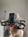 A hand is holding a small drone equipped with a camera at the front. The drone has four propellers, and the camera is enclosed within a black casing. The background is softly blurred, emphasizing the focus on the drone.