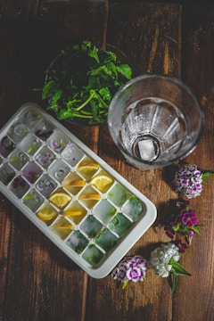 A wooden surface features a tray of ice cubes, each compartment containing colorful additions like lemon slices and herbs. Beside it is an empty glass and a small bouquet of fresh flowers. In the background, there are green leaves placed in a pile.