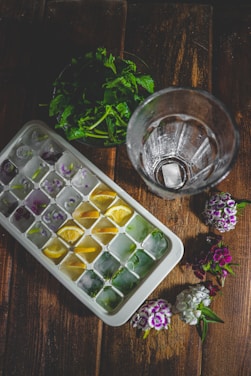 A wooden surface features a tray of ice cubes, each compartment containing colorful additions like lemon slices and herbs. Beside it is an empty glass and a small bouquet of fresh flowers. In the background, there are green leaves placed in a pile.