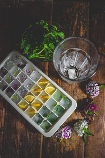 A wooden surface features a tray of ice cubes, each compartment containing colorful additions like lemon slices and herbs. Beside it is an empty glass and a small bouquet of fresh flowers. In the background, there are green leaves placed in a pile.