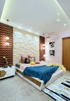 A modern bedroom with geometric wall panels and vibrant decor. The bed is adorned with colorful pillows and a blue blanket. Two pendant lights hang above each bedside table, enhancing the room's ambiance. Black and white photographs hang on the wall, and a textured rug lies on the floor.