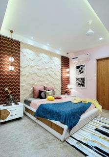 A modern bedroom with geometric wall panels and vibrant decor. The bed is adorned with colorful pillows and a blue blanket. Two pendant lights hang above each bedside table, enhancing the room's ambiance. Black and white photographs hang on the wall, and a textured rug lies on the floor.